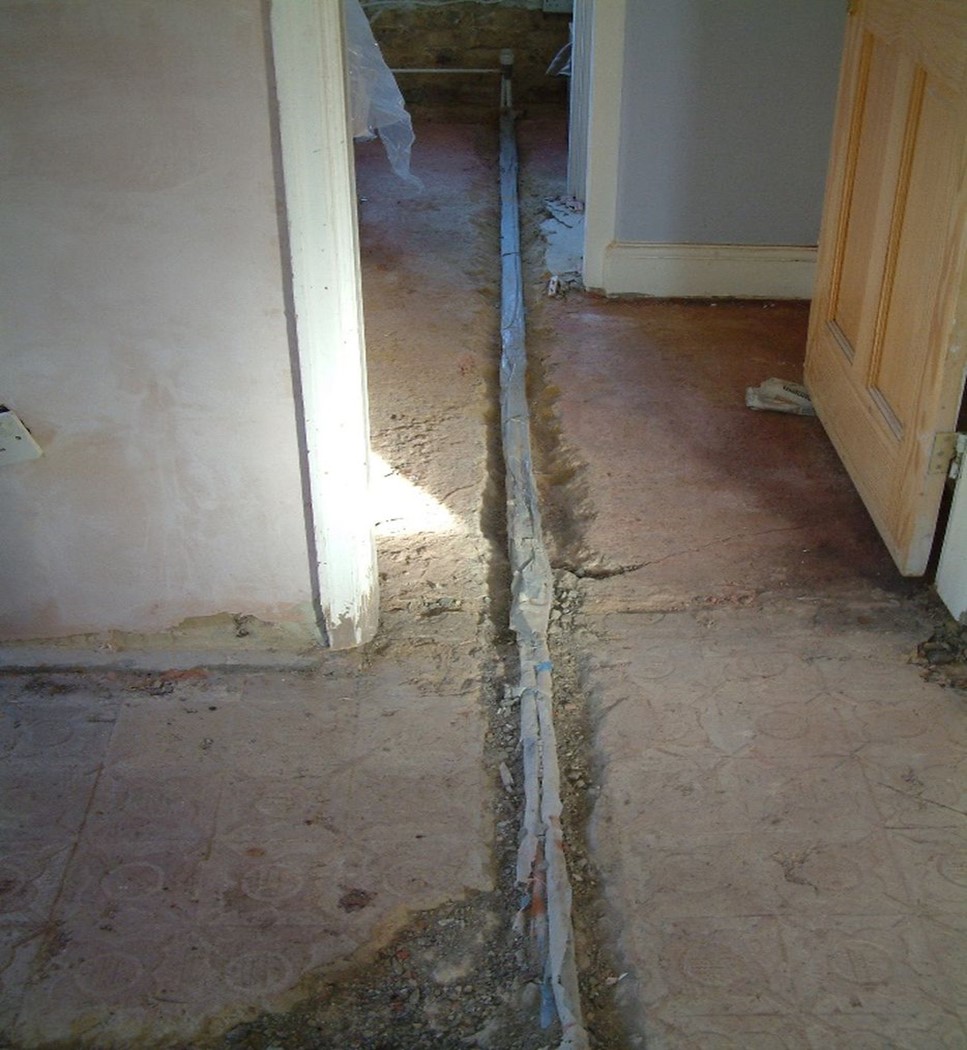 A photograph of a concrete floor inside a house. The floor has been dug up to reveal a corroded copper pipe running in a straight line very close to the surface.